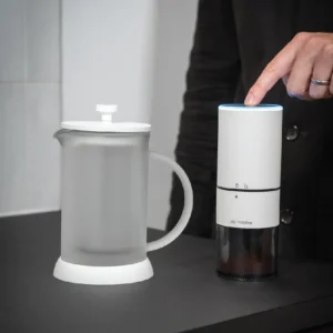 User pressing the power button on white Groove Compact Electric Grinder next to a French press