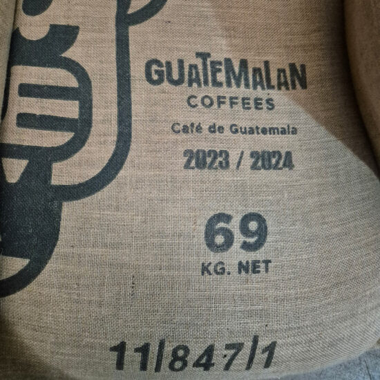 A burlap coffee sack labeled with 'Guatemalan Coffees' and details like crop year 2023/2024, weighing 69 kg net, from Guatemala